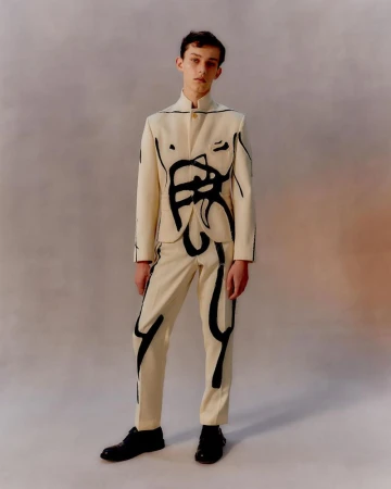 Adult male model posing in a studio wearing a cream-colored pantsuit with black abstract graphic art, and black patent leather shoes.