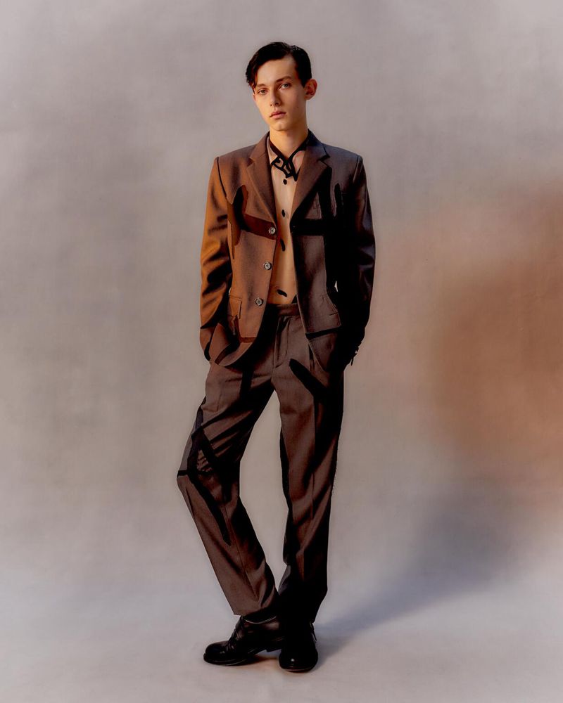A young adult stands in a studio, wearing a gray suit with black accents, black shoes, and a light-colored shirt and tie with dark pattern.