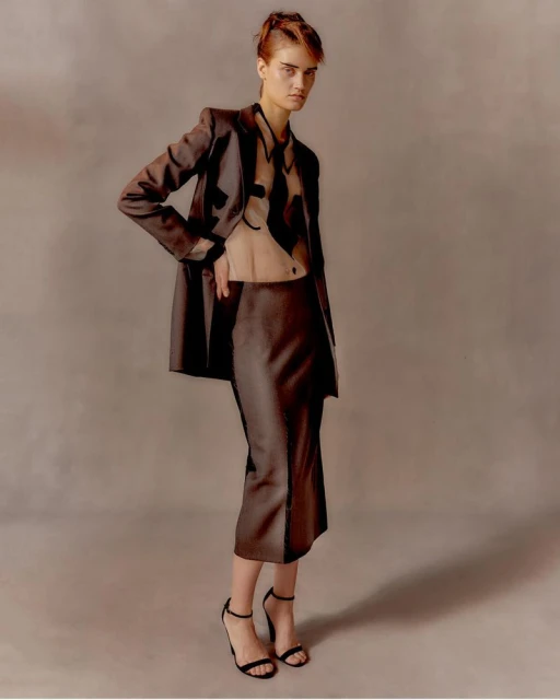 A slender fashion model poses indoors wearing a brown skirt suit and black strappy high heels in a studio environment with muted background.