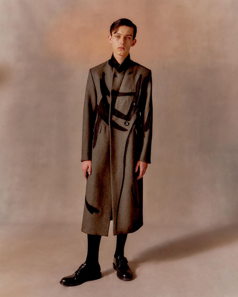 A young man poses in a long, patterned coat with black socks and dress shoes against a gradient neutral background in a studio shot.