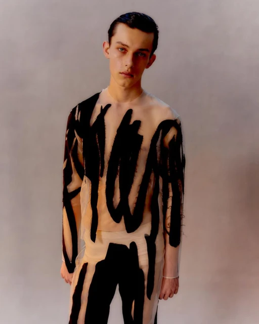 Portrait of a young adult with dark hair, wearing a sheer top and pants with black brushstroke pattern, against a neutral backdrop.