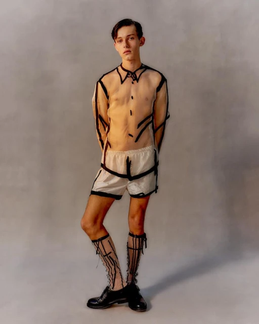 Full-length studio portrait of a young adult male in a sheer shirt and shorts, with painted black lines, striking a pose against a grey backdrop.
