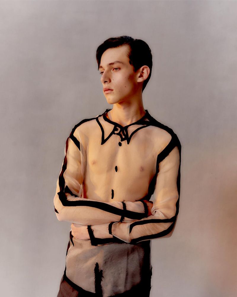 Portrait of a young adult male in a sheer top with black outline, arms crossed, gazing left, studio lighting, and neutral background.