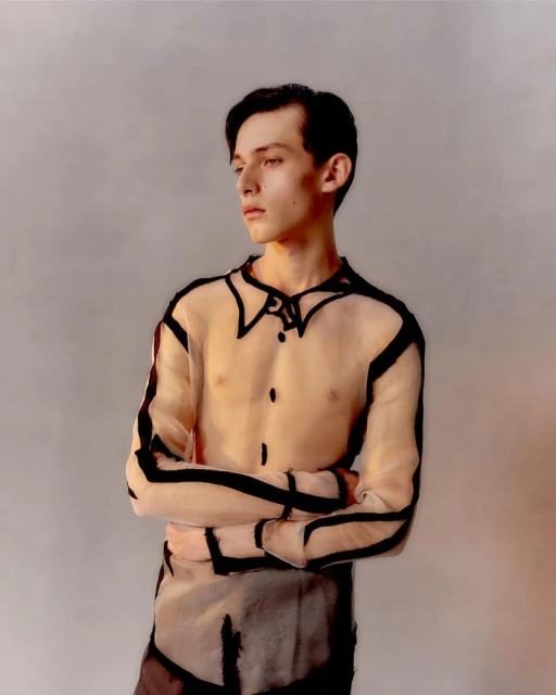 Portrait of a young adult male in a sheer top with black outline, arms crossed, gazing left, studio lighting, and neutral background.