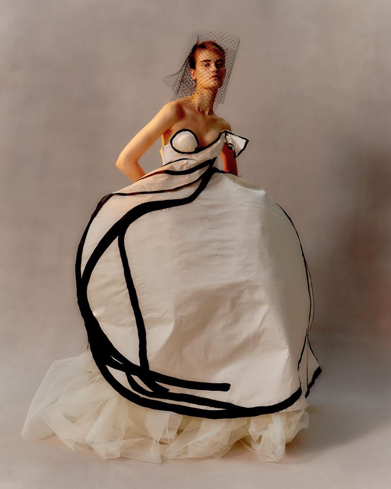Woman in a large white and black formal gown with a veil stands in a photo studio in a fashion portrait. The style is editorial.