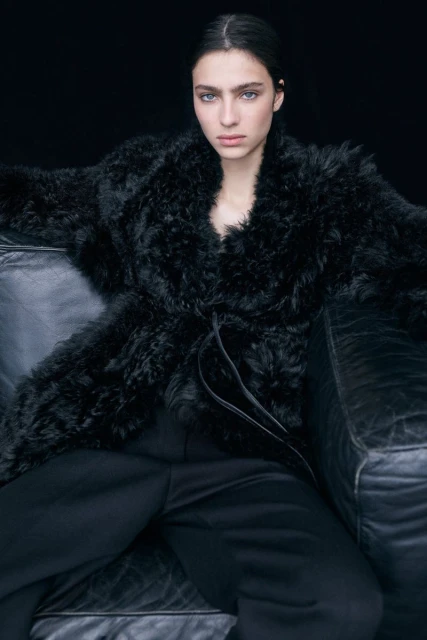 A striking studio shot showcases a woman with fair skin and dark hair, posing in a black fur coat and pants, seated on a black couch against a dark backdrop.