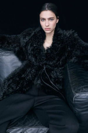 A striking studio shot showcases a woman with fair skin and dark hair, posing in a black fur coat and pants, seated on a black couch against a dark backdrop.