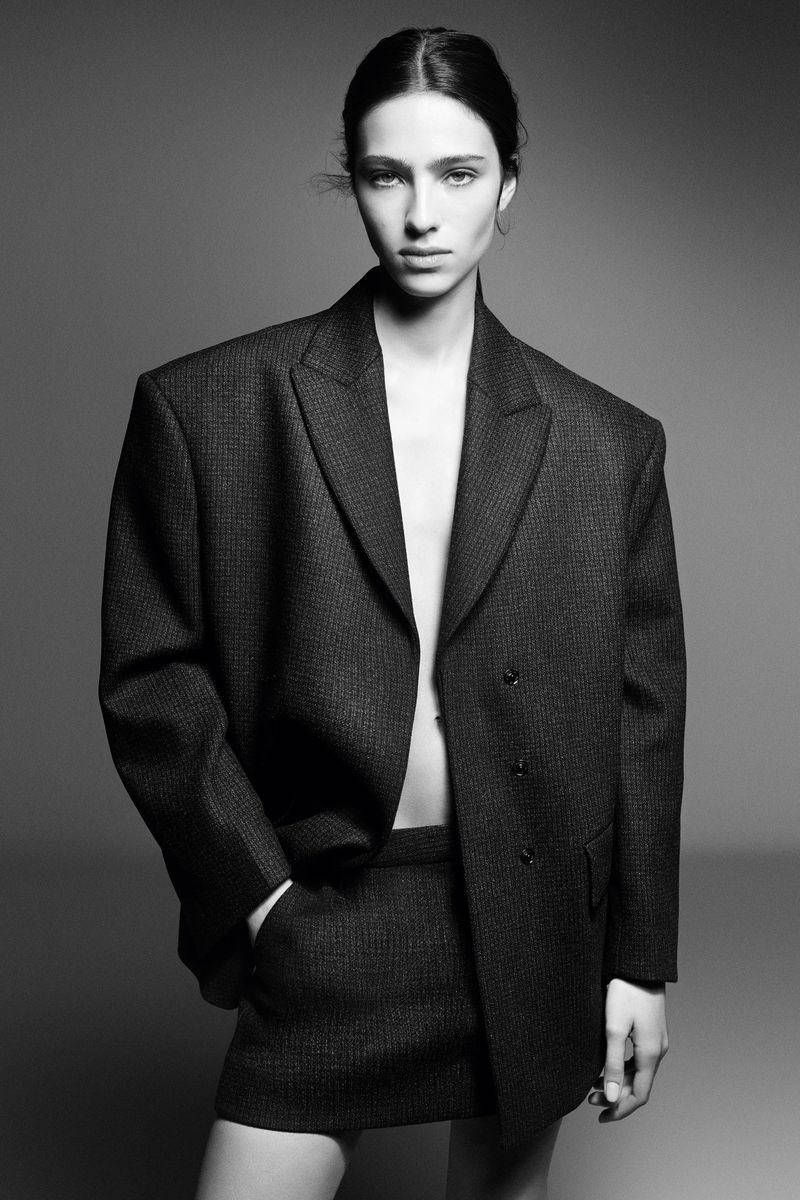 Model Loli Bahia posing in an oversized dark grey tweed blazer and matching mini skirt against a neutral studio backdrop.