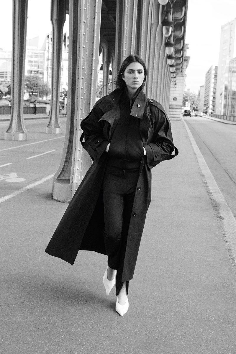 Loli Bahia walking along an architectural bridge in an oversized dark coat and white pointed heels for H&M Studio.
