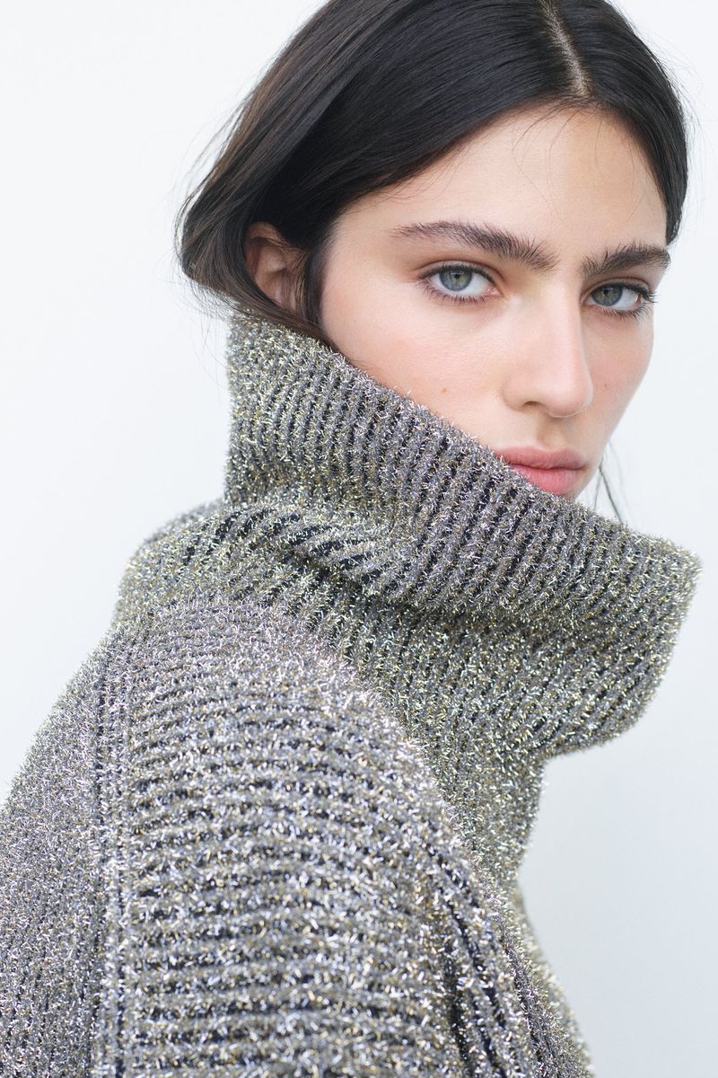 Model Loli Bahia looking over her shoulder wearing a textured metallic ribbed turtleneck sweater against a plain white background.