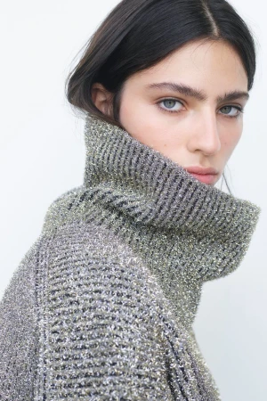 Model Loli Bahia looking over her shoulder wearing a textured metallic ribbed turtleneck sweater against a plain white background.