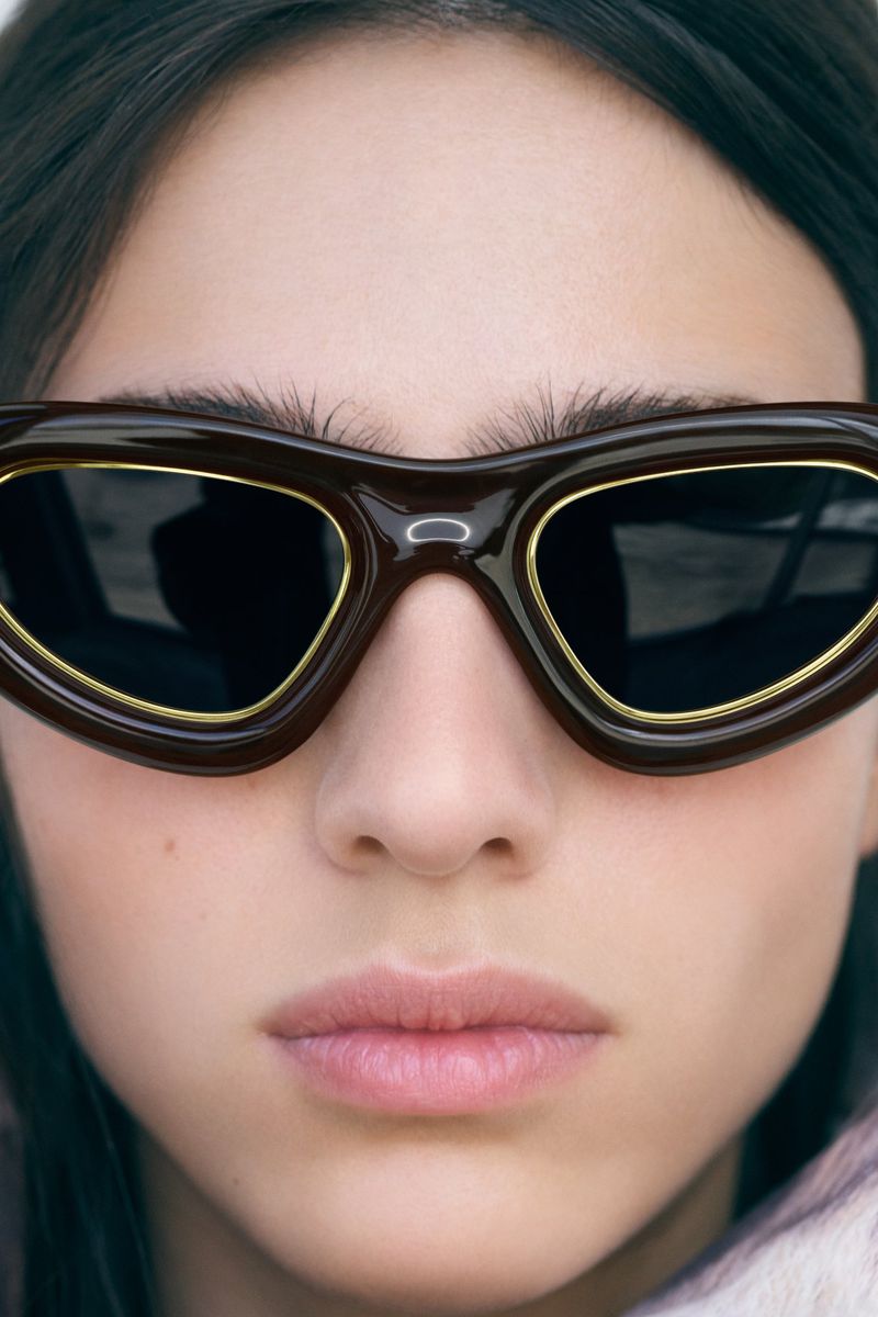 Close-up portrait of a fair-skinned woman wearing dark brown sunglasses with gold trim, showcasing fashion and beauty elements.