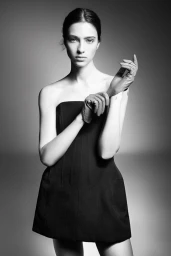 Black and white studio portrait of model Loli Bahia wearing a structured strapless black mini dress while adjusting gloves.