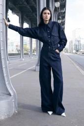 Model Loli Bahia wearing an oversized dark blue denim shirt and wide-leg trousers while posing against a steel bridge pillar.