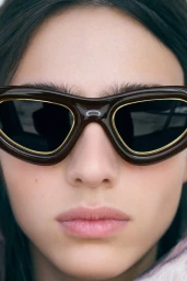 Close-up portrait of model Loli Bahia wearing glossy dark brown sunglasses with gold-rimmed lenses.