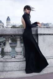 Model Loli Bahia in a floor-length backless black dress posing along a stone bridge balustrade with city buildings behind.