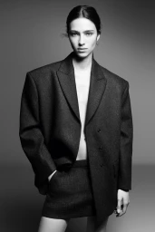 Model Loli Bahia posing in an oversized dark grey tweed blazer and matching mini skirt against a neutral studio backdrop.