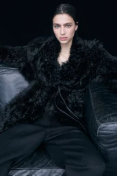 Model Loli Bahia seated on a dark leather couch wearing a black faux fur coat for the H&M Studio F/W 24 lookbook.
