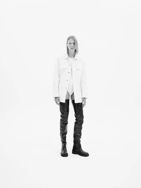 A single adult female stands facing forward in a white coat with black, over-the-knee boots against a solid, bright white background.