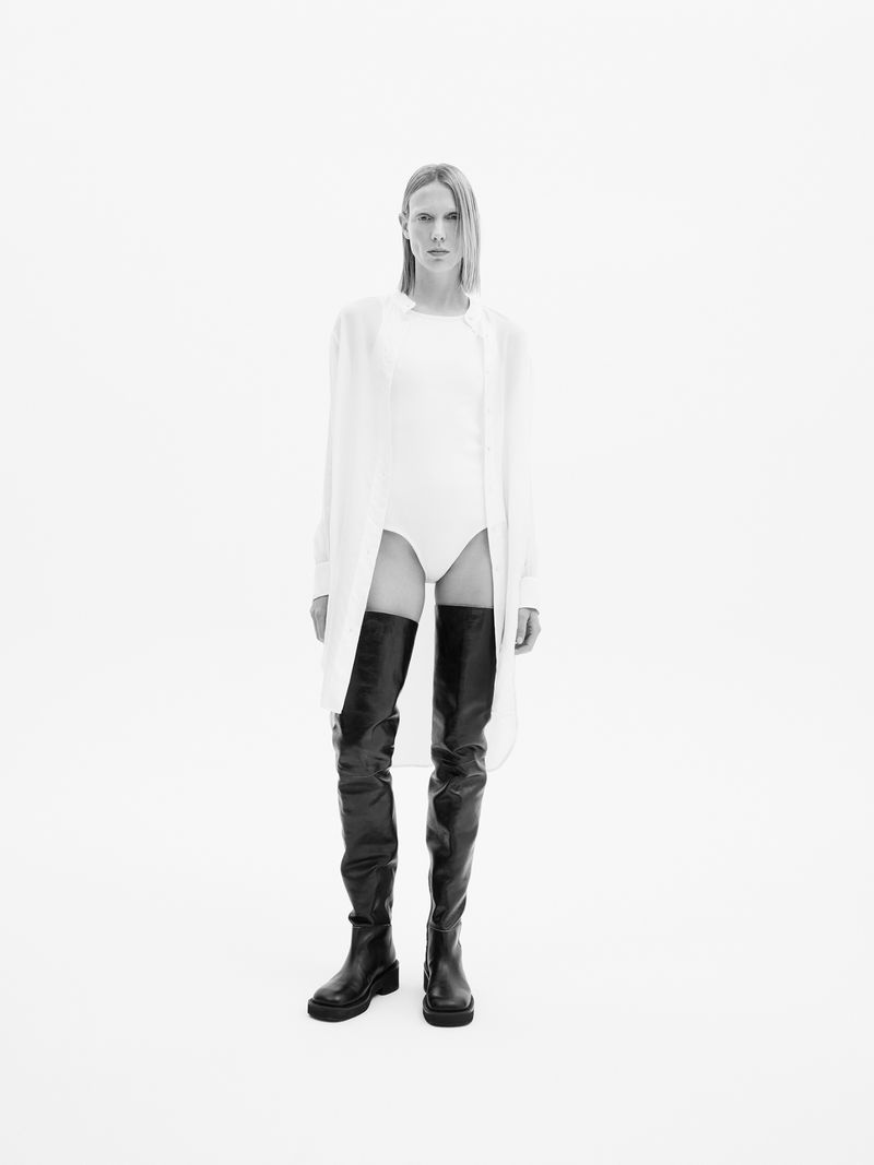 Model poses in a white bodysuit, oversized shirt, and thigh-high black boots against a white background in a studio fashion shot.