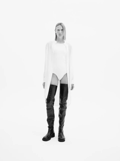 Model poses in a white bodysuit, oversized shirt, and thigh-high black boots against a white background in a studio fashion shot.