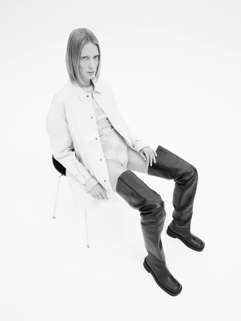 Black and white shot of a model in a shirt dress and thigh-high boots, seated against a stark white backdrop, creating a high-contrast fashion statement.