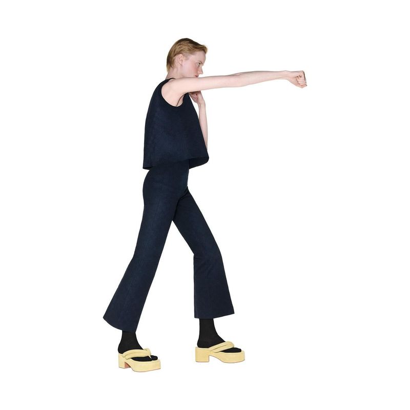 Woman in dark blue two-piece outfit stands in a boxing pose with platform sandals against a bright white background in a studio shot.