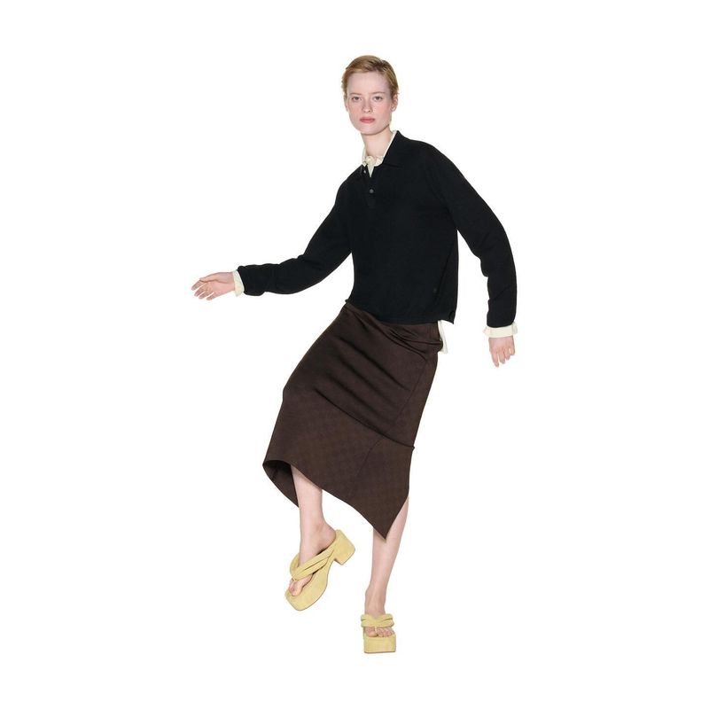 Woman in black sweater and brown skirt stands with one leg raised against a white background, showcasing a fashion forward pose and style.