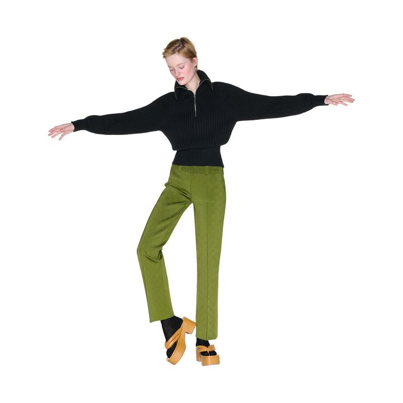 A young woman in a black quarter-zip sweater, green trousers, and platform sandals is posing in a studio, arms extended out.