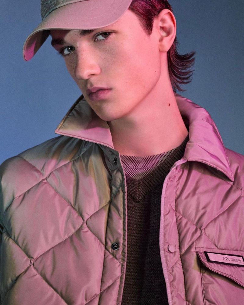 Portrait of a young man with a baseball cap, wearing a quilted jacket and sweater. Soft lighting and a blue background.