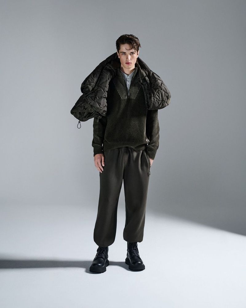 A young adult male is posing in a studio, wearing dark green sweatpants and a matching sweater, accessorized with black boots and a puffer jacket.