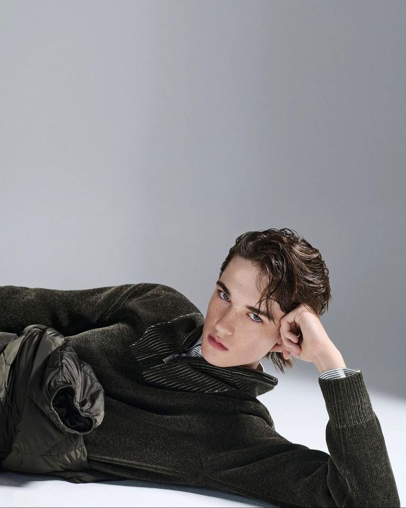 A young man lies in a relaxed pose wearing a dark green sweater and olive-green vest against a neutral studio backdrop.