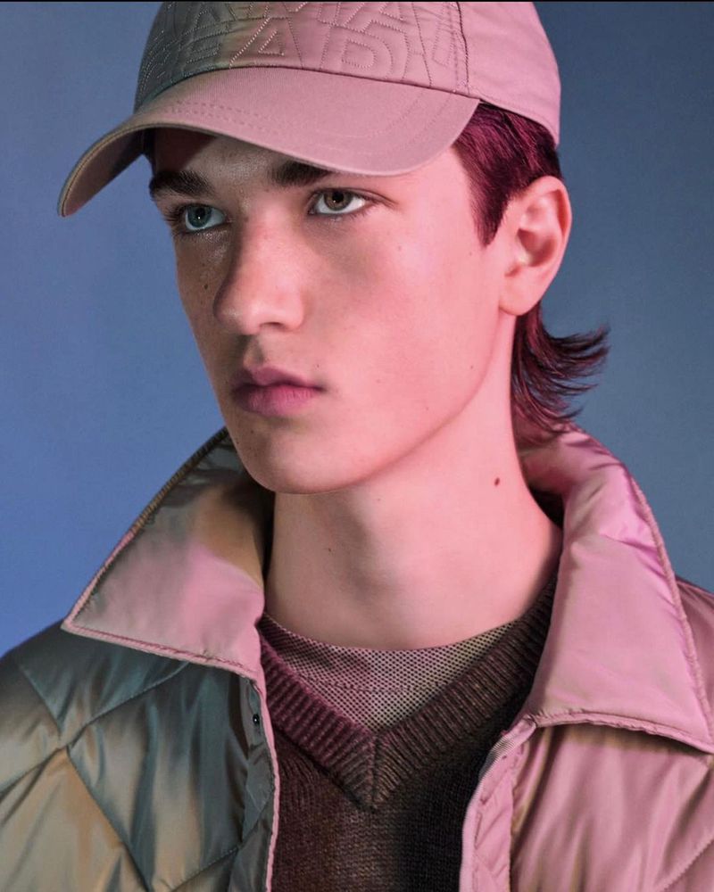 Close-up portrait of a young adult male wearing a baseball cap and quilted jacket, in a studio setting with a blue gradient background.