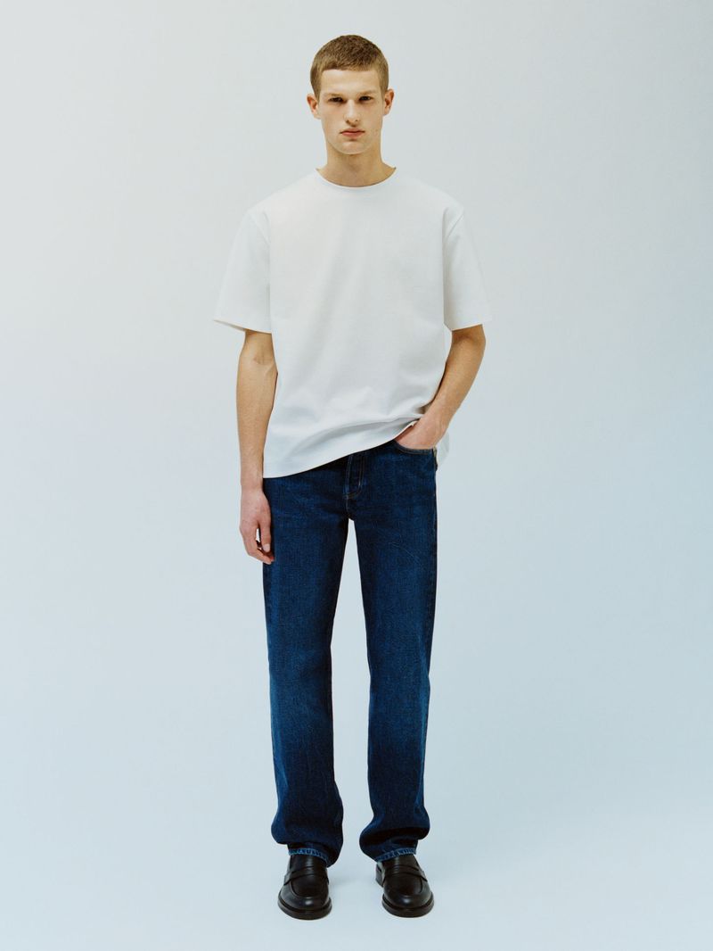 Model Teun van Stein wearing a white crewneck t-shirt, dark blue jeans, and black loafers against a light seamless studio backdrop.