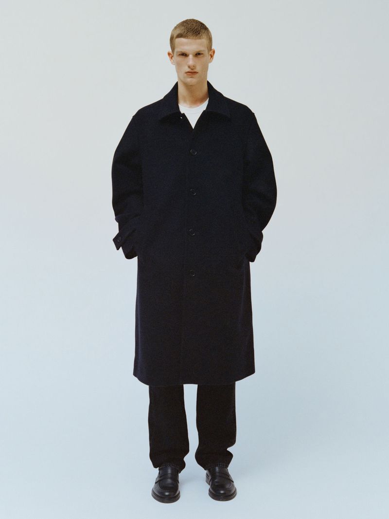 Full-length shot of a fair-skinned adult male in an overcoat, trousers, and dress shoes, posing in a bright photo studio against a light blue backdrop.