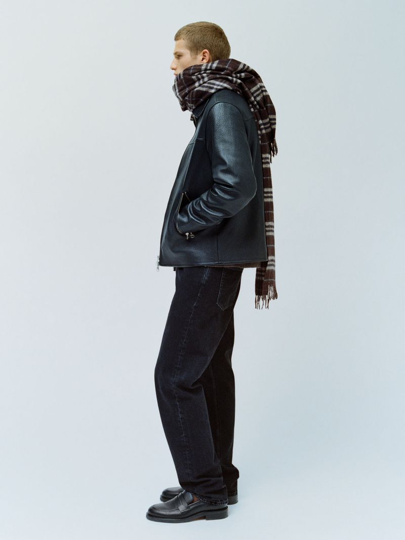Side profile of a man wearing a black leather jacket, dark trousers, and a plaid scarf against a seamless light grey studio backdrop.