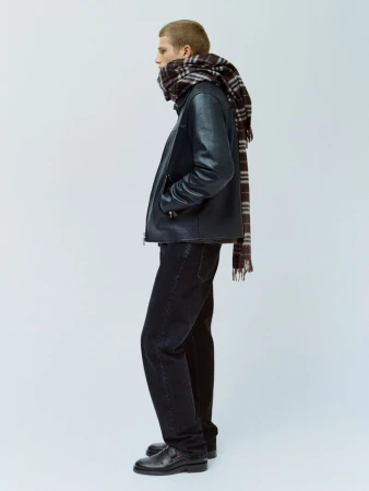 Side profile of a man wearing a black leather jacket, dark trousers, and a plaid scarf against a seamless light grey studio backdrop.