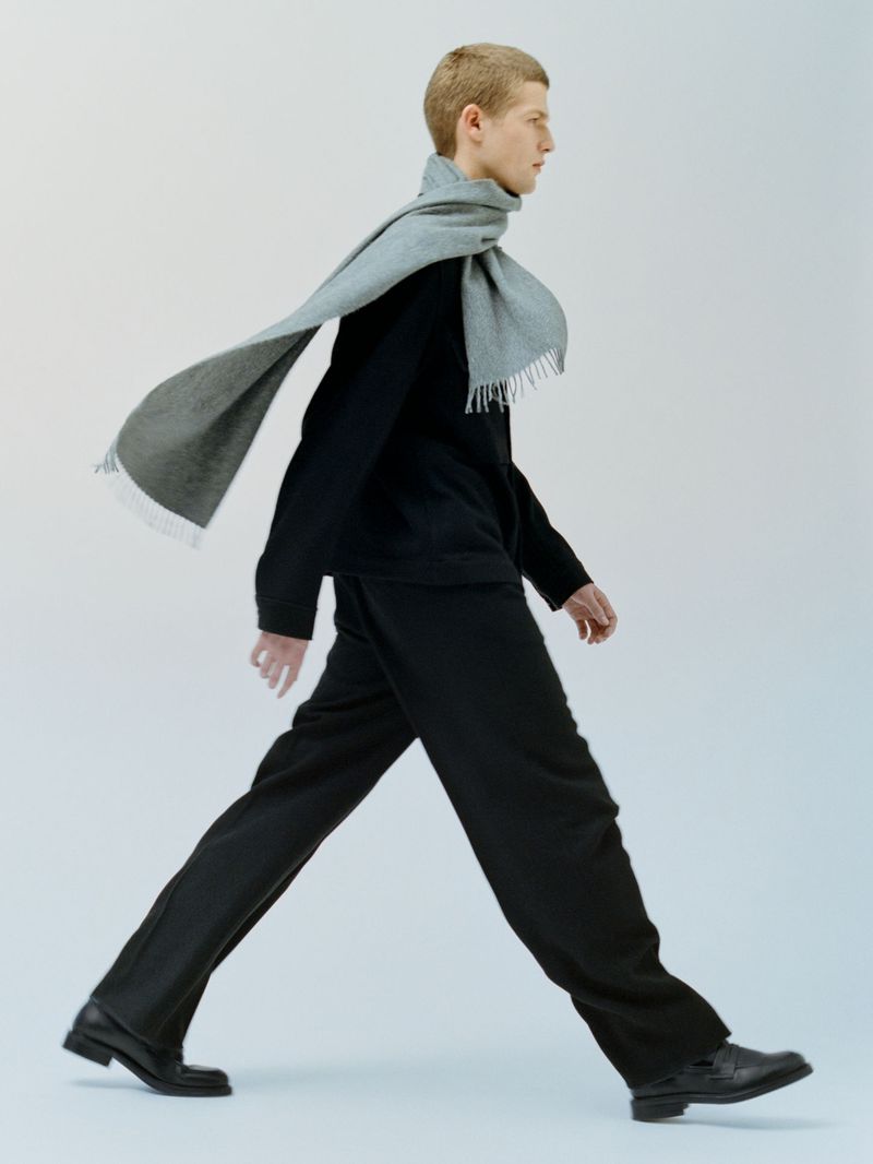 Side view of a fair-skinned male model walking in a black outfit and a gray scarf against a plain white backdrop in a studio shot.