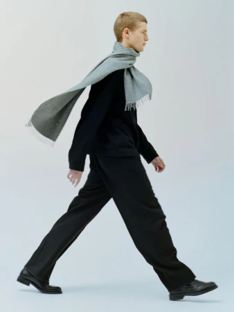 Side view of a fair-skinned male model walking in a black outfit and a gray scarf against a plain white backdrop in a studio shot.
