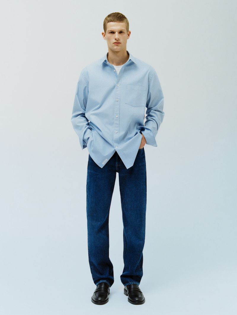 A young adult man in a blue collared shirt and dark wash jeans stands in a brightly lit photo studio with his hands in his pockets.