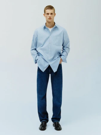 A young adult man in a blue collared shirt and dark wash jeans stands in a brightly lit photo studio with his hands in his pockets.