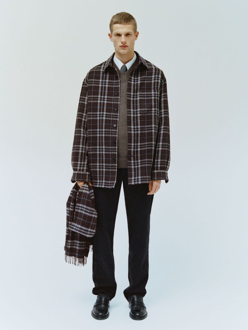 Studio shot of a male model wearing a plaid shirt and dark trousers, holding a matching plaid scarf. White backdrop.