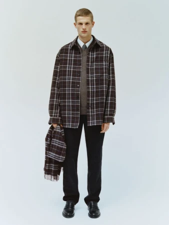 Studio shot of a male model wearing a plaid shirt and dark trousers, holding a matching plaid scarf. White backdrop.