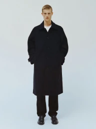 Model Teun van Stein wearing a long black coat, dark trousers, and leather shoes against a pale studio backdrop for Arket.