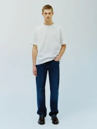 Model Teun van Stein wearing a white crewneck t-shirt, dark blue jeans, and black loafers against a light seamless studio backdrop.