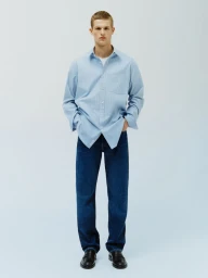 Model Teun van Stein standing in an oversized light blue button-down shirt and dark blue denim jeans against a pale blue background.
