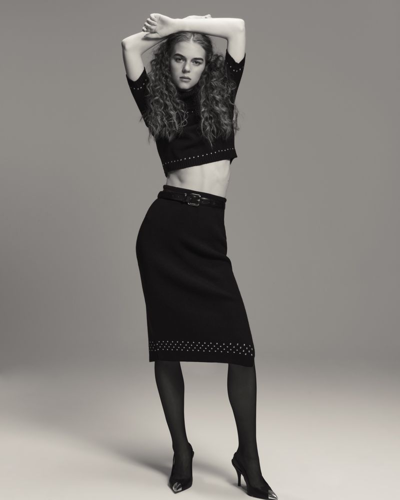 A woman with curly hair poses in a cropped sweater, black skirt, black tights, and high heels in a studio environment.