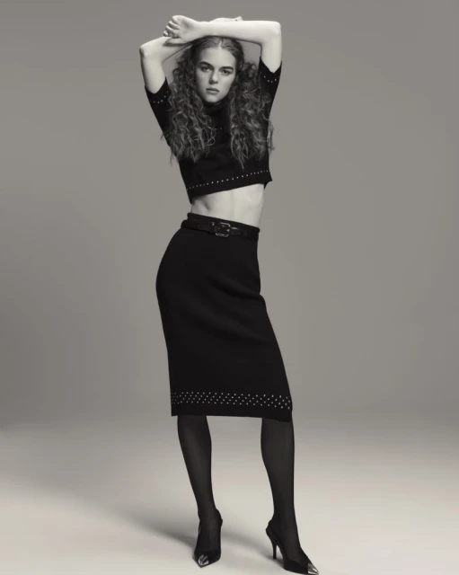 A woman with curly hair poses in a cropped sweater, black skirt, black tights, and high heels in a studio environment.