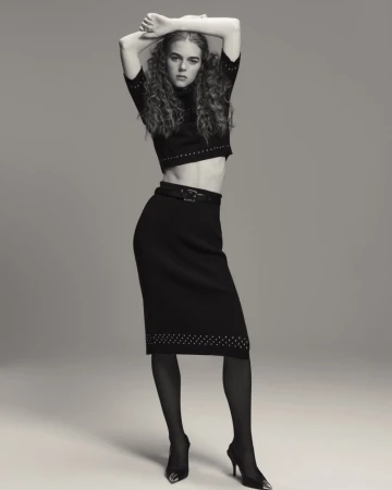 A woman with curly hair poses in a cropped sweater, black skirt, black tights, and high heels in a studio environment.
