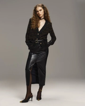 Model Tilda Jönsson posing in a black knit cardigan and a black leather slit midi skirt against a neutral grey studio backdrop.